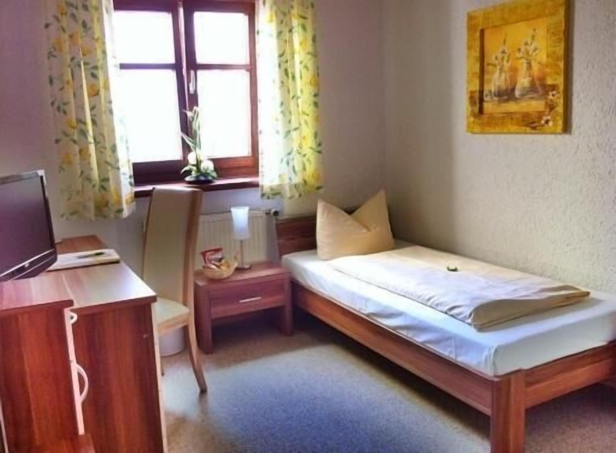 A small, neatly arranged bedroom with a single bed, a desk with a computer, and a window with curtains.
