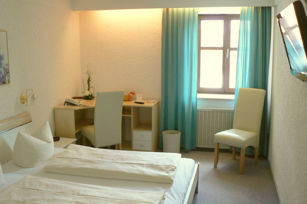A hotel room with a bed, a desk, a chair, a radiator, and a window with curtains.