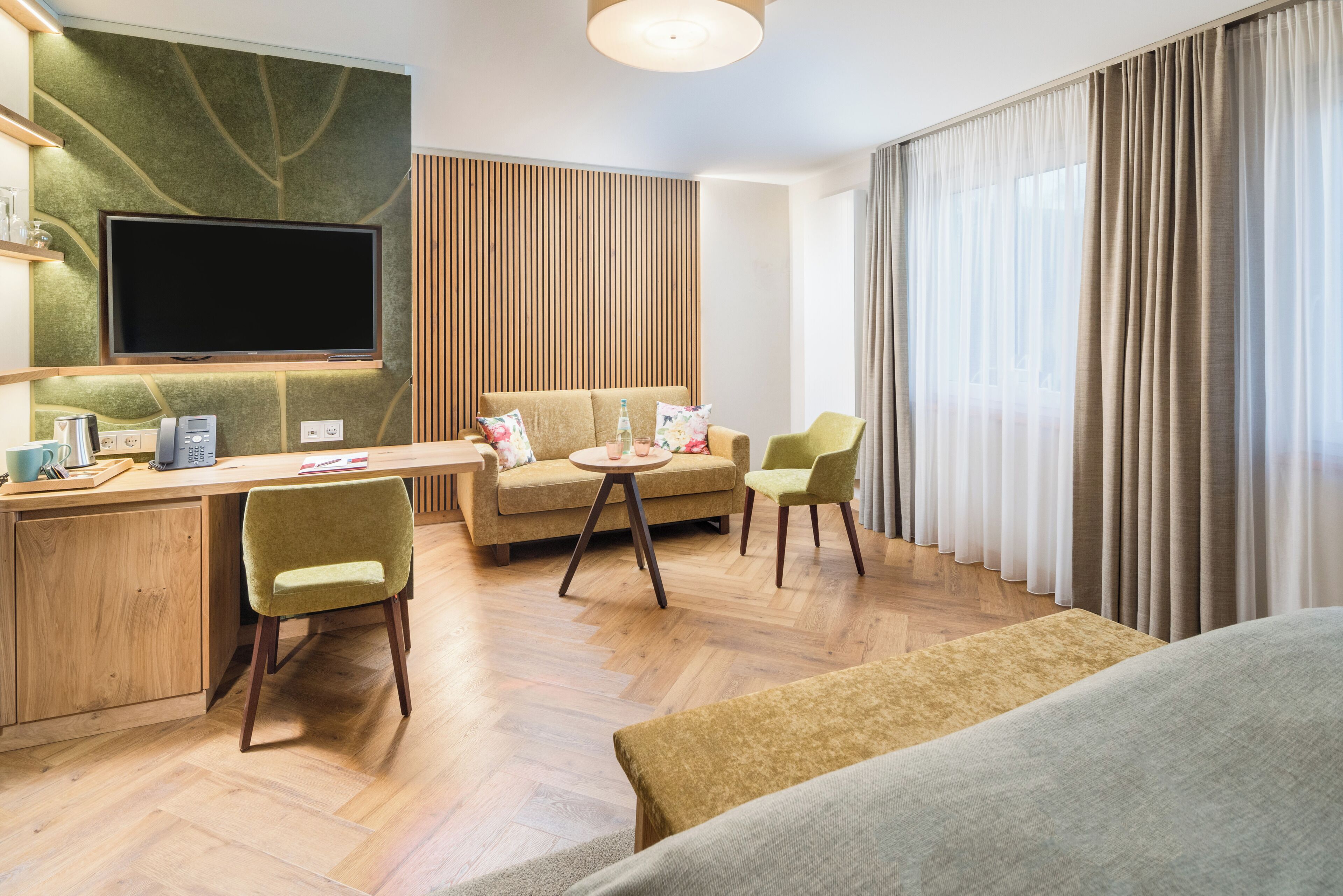 A modern hotel room with a flat-screen TV, a wooden desk, a sofa, two chairs, and a large window with curtains.