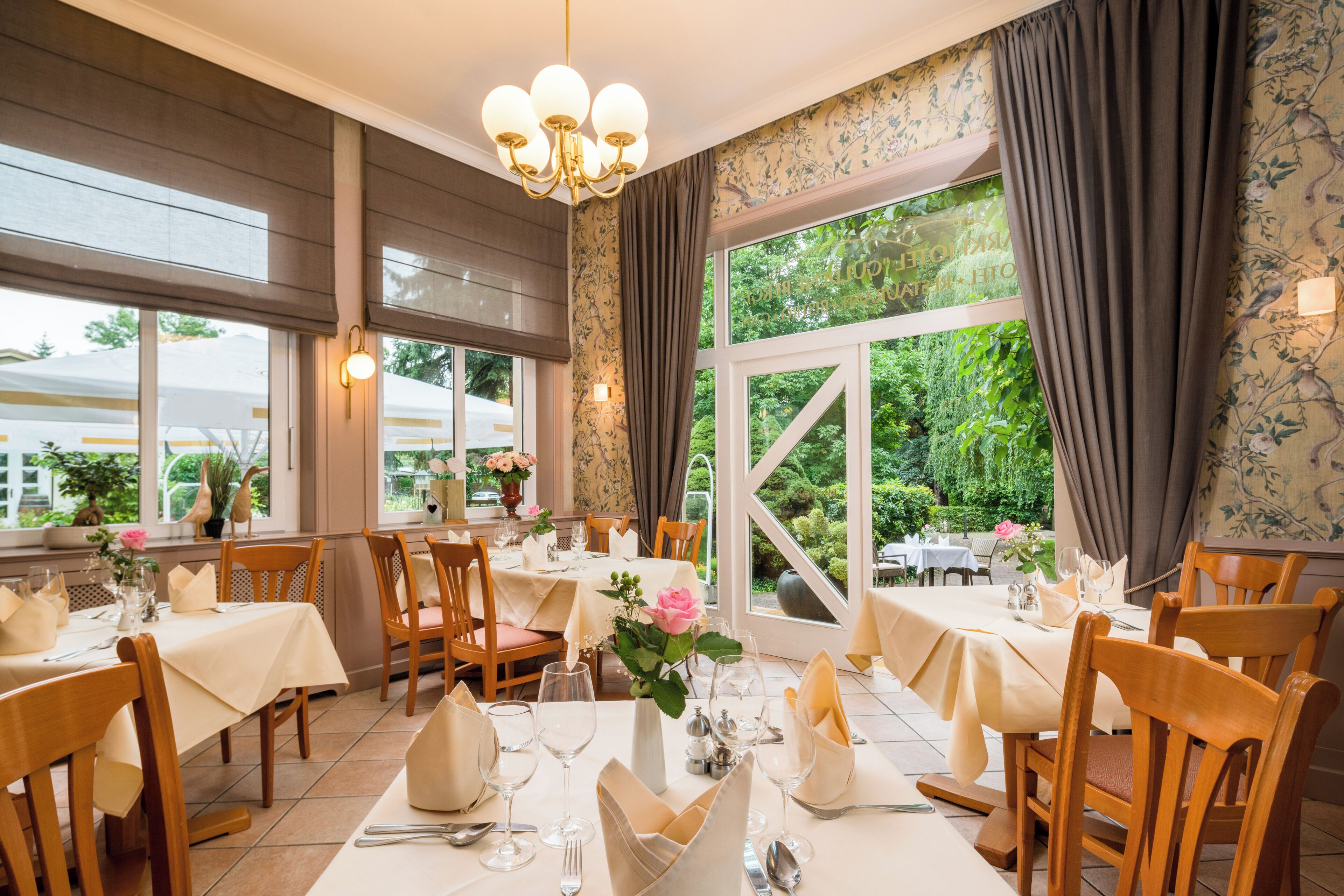 Ein Restaurant mit mehreren Esstischen, großen Fenstern mit Vorhängen und einer Kristallleuchtre.