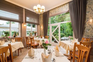 Un restaurant avec de nombreuses tables dressées pour le service, de grandes fenêtres avec des rideaux et un lustre.