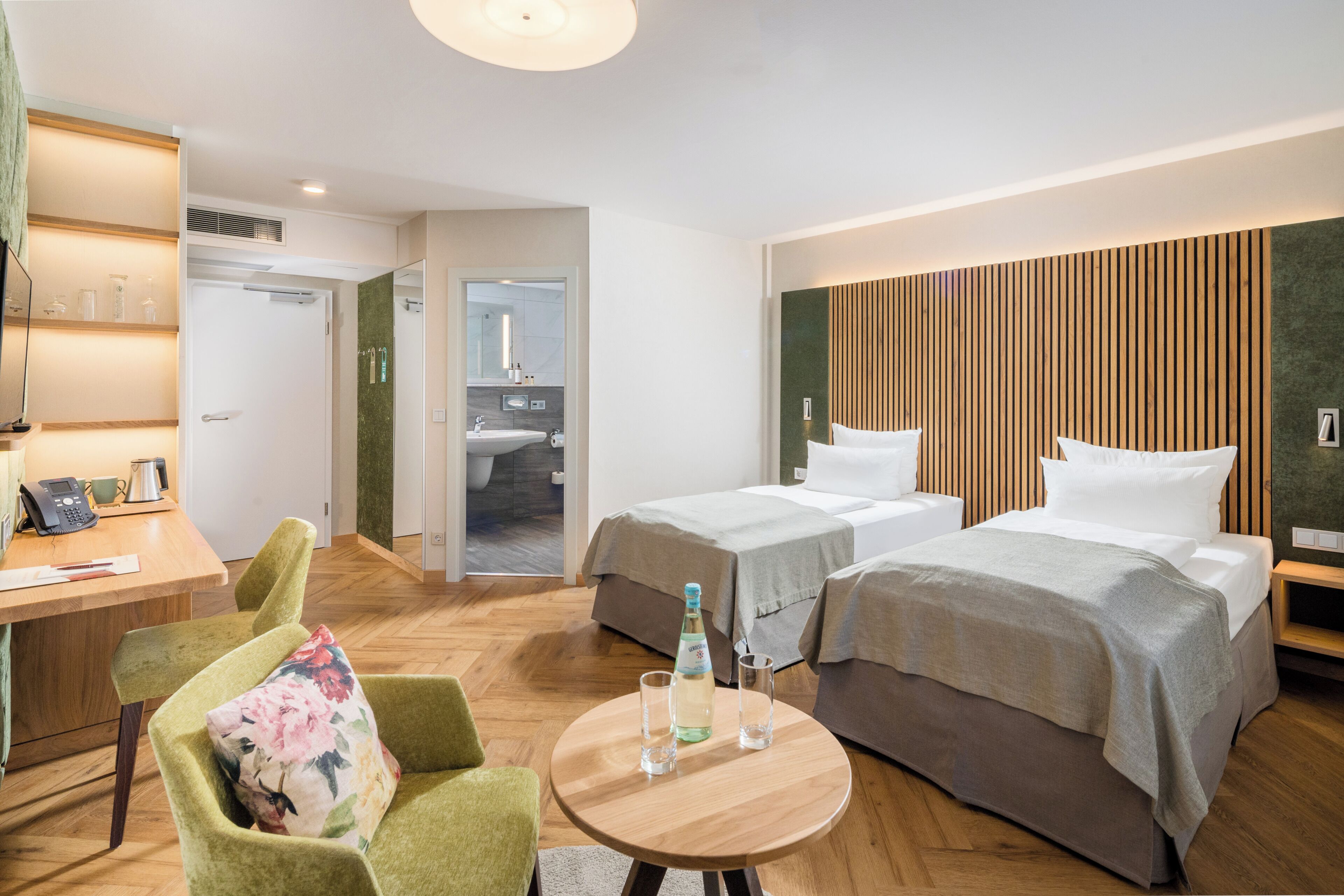 A hotel room with two beds, a desk with a phone and a kettle, and a chair with a floral cushion.
