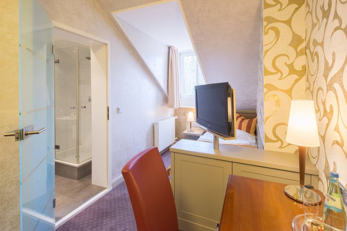 Habitación de hotel con cabina de ducha de vidrio, cama con televisor, escritorio con lámpara y una botella de agua.