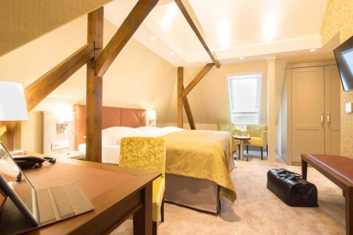 A hotel room with a bed, a desk with a laptop, a suitcase, and a TV on the wall.