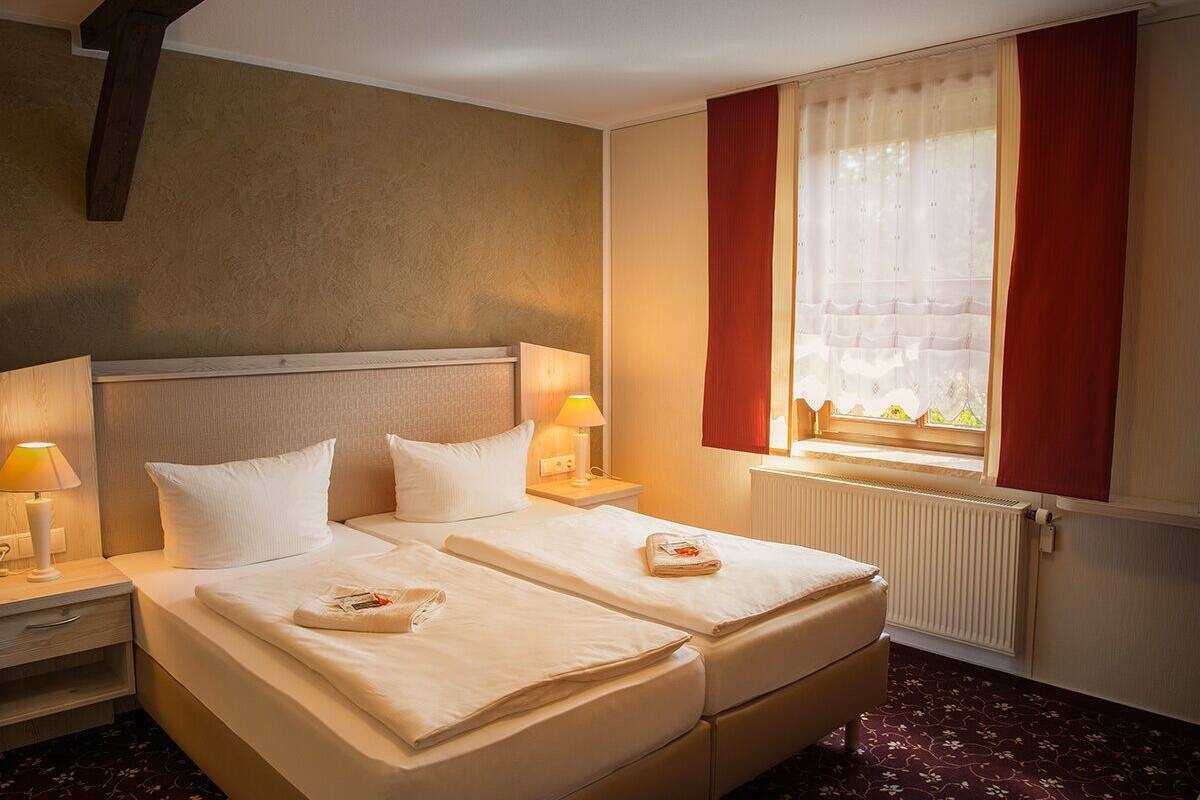 A hotel room with two beds, a window with red curtains, a radiator, and bedside lamps.