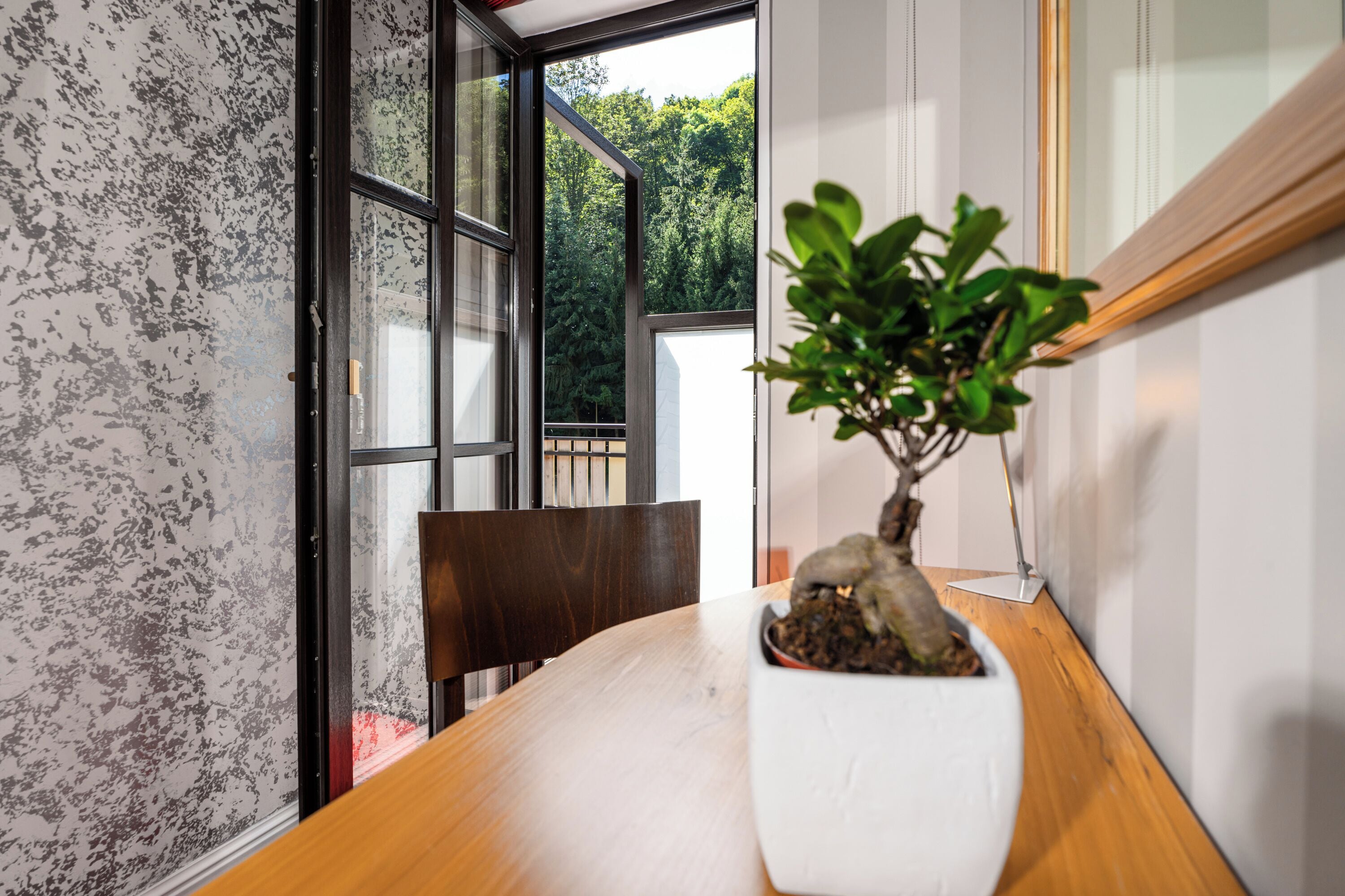 A room with a wooden table, a white potted plant, a chair, and a window with a view of trees.