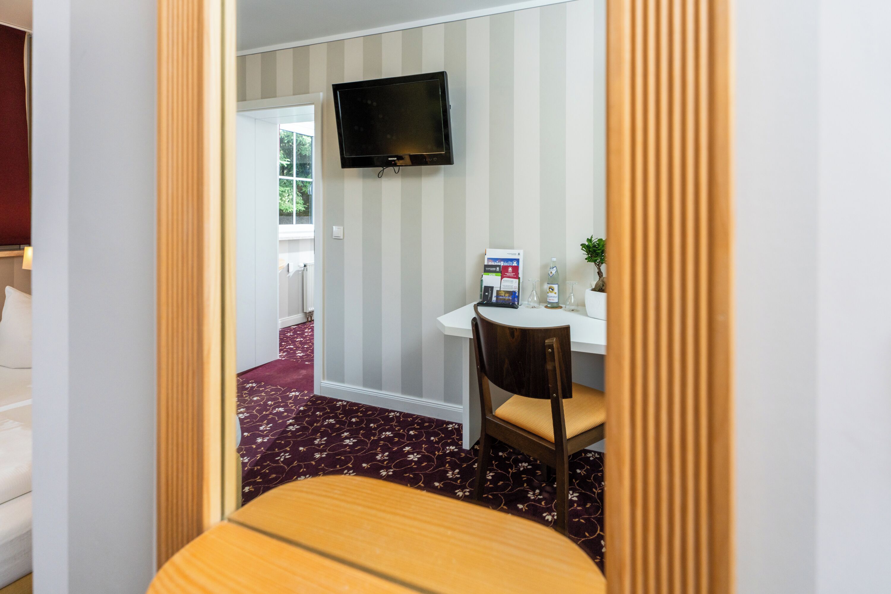 Ein Zimmer mit an der Wand montiertem Fernseher, einem Schreibtisch mit Stuhl, einer kleinen Pflanze und einem Fenster.