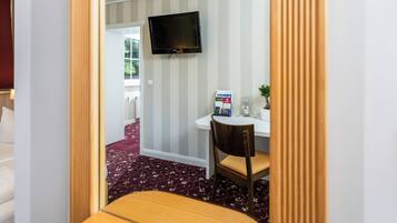 Ein Zimmer mit an der Wand montiertem Fernseher, einem Schreibtisch mit Stuhl, einer kleinen Pflanze und einem Fenster.