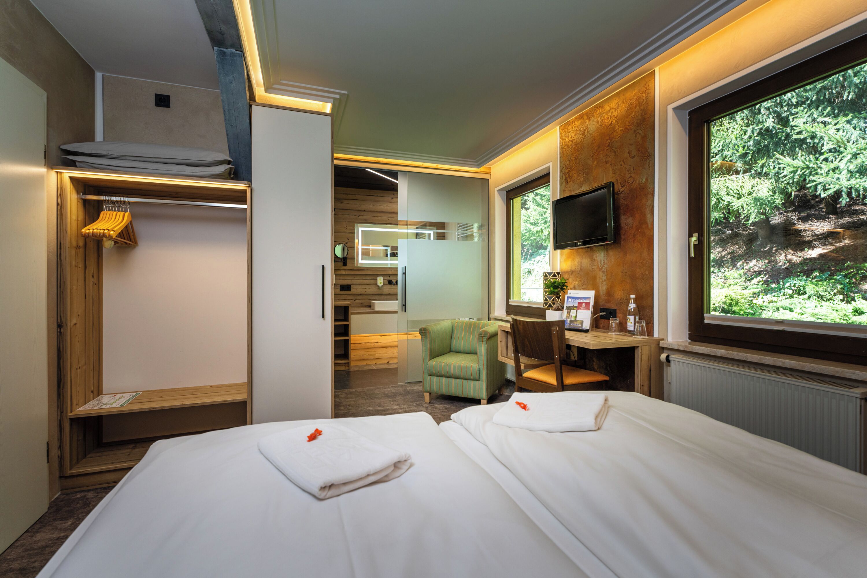 A hotel room with two beds, a TV, a desk with a chair, a wardrobe, and a large window with a view of trees.