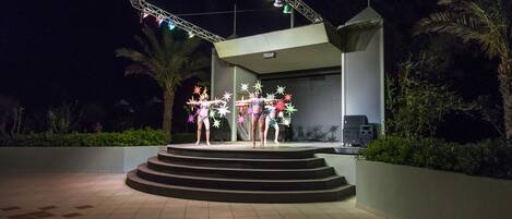 A stage with a metal truss and lights, surrounded by palm trees and a building with steps leading up to it.