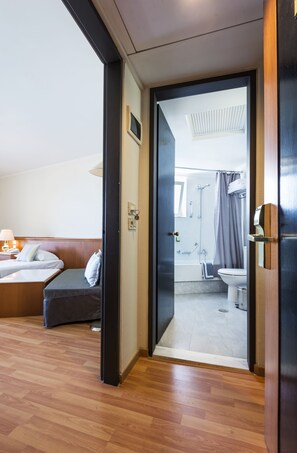A hotel room with a wooden floor, a bed with pillows, a bedside table with a lamp, and a bathroom with a shower, toilet, and bathtub.