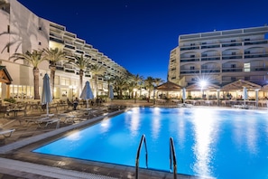 A swimming pool with lounge chairs and umbrellas, surrounded by palm trees and a building with balconies.