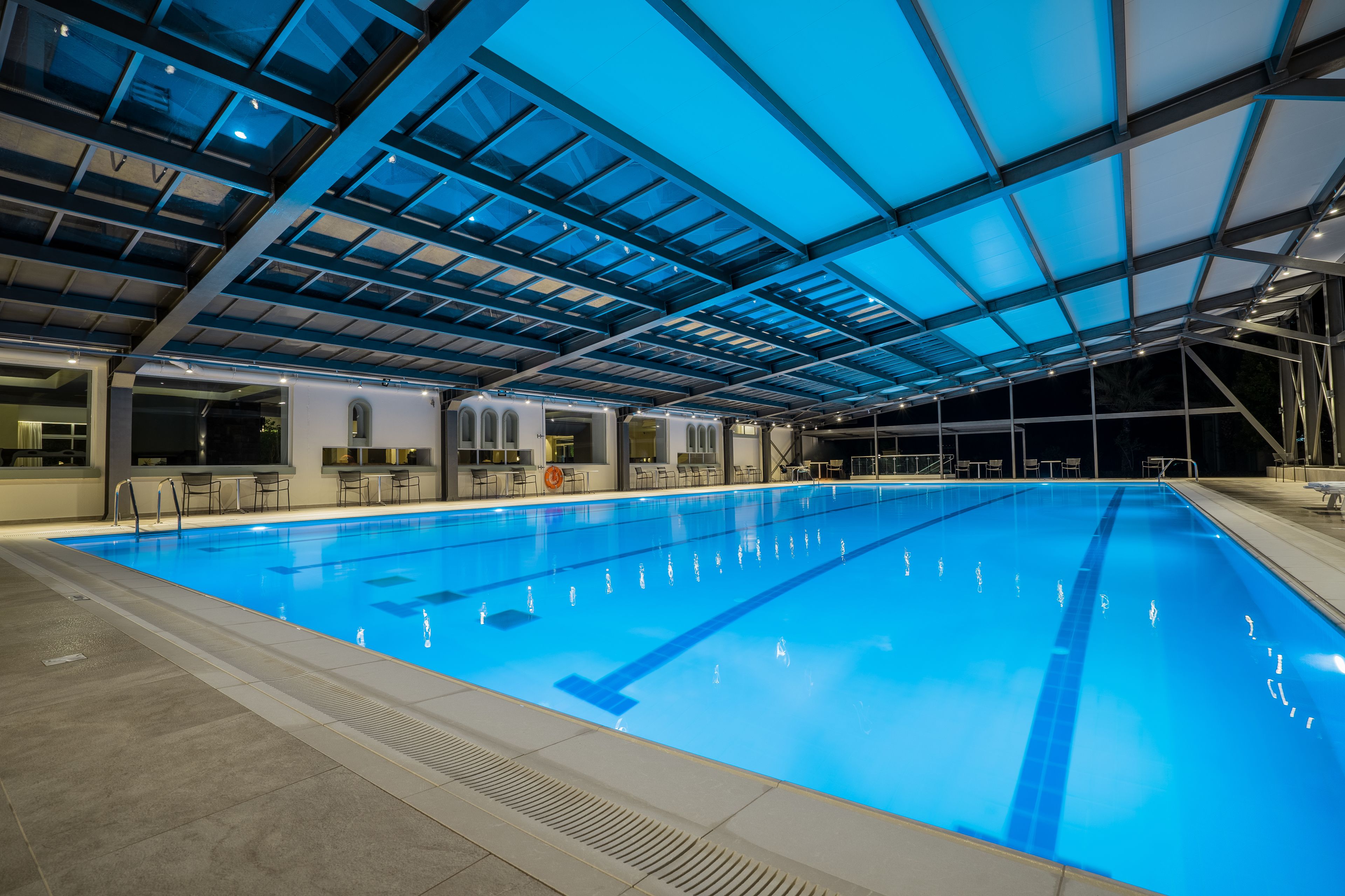 An indoor swimming pool with a glass ceiling, tiled floor, and seating area along the pool's edge.