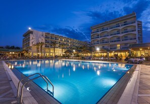 A swimming pool with a poolside area, a multi-story hotel building, and palm trees.