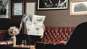 A person reading a newspaper in a room with a brown leather couch, framed pictures on the wall, and a vase with dried flowers on a side table.