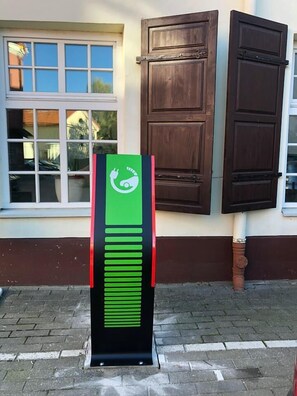 A green and black electric vehicle charging station with a plug symbol, located on a paved sidewalk next to a building with brown shutters and a window. - Shakespeare Boutique Hotel (Vilnius)