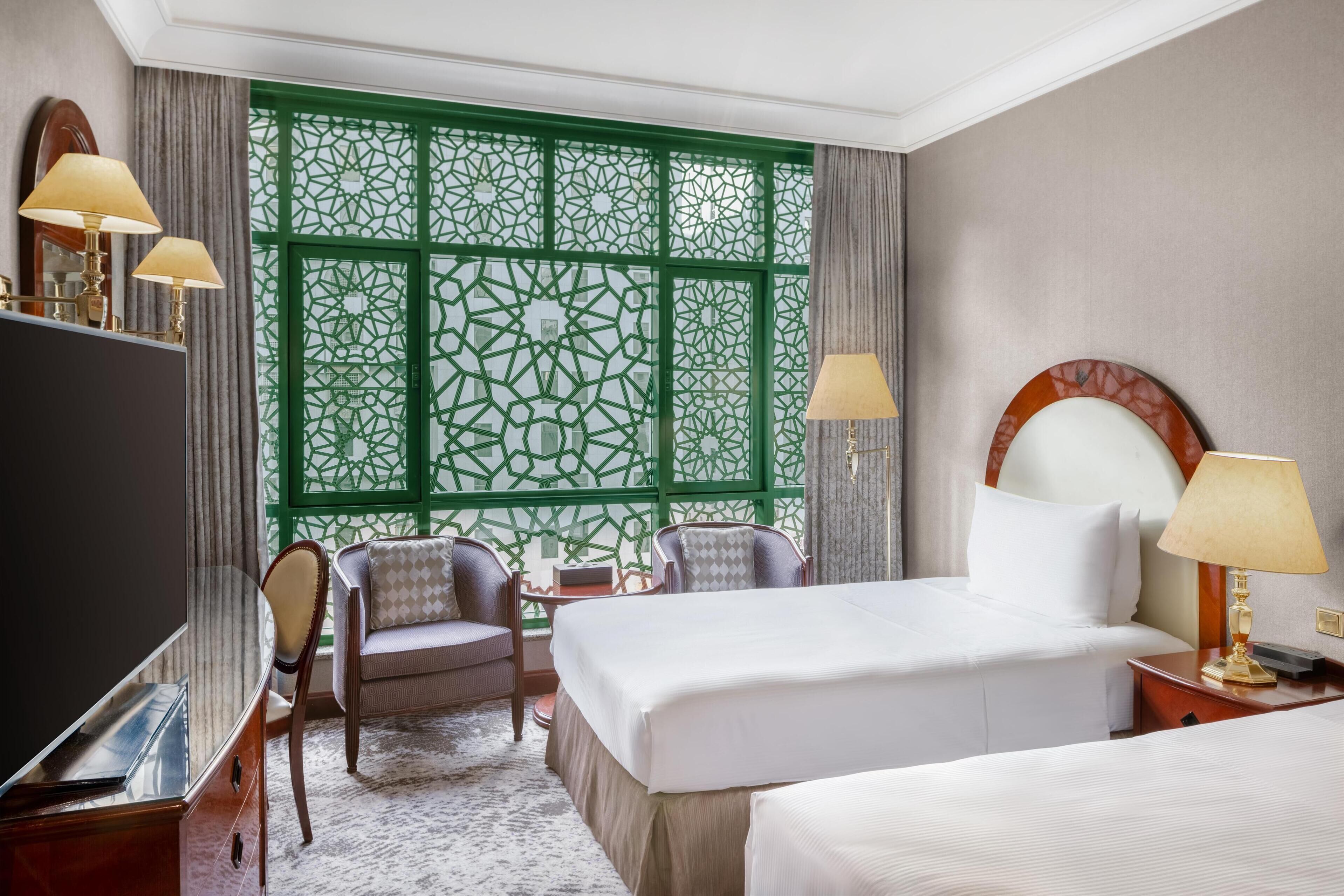 A hotel room with a large bed, a desk with a TV, two chairs, a small table, and a decorative window with green patterns.