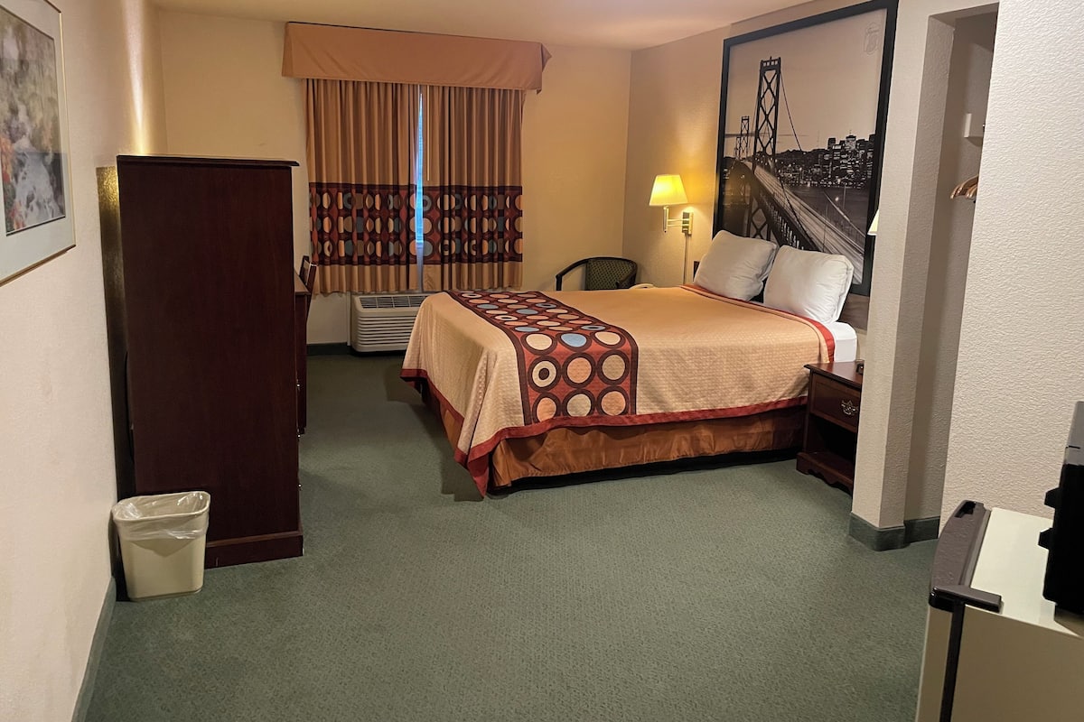 A hotel room with a bed, a desk, a chair, a waste bin, a lamp, and a painting of a bridge.