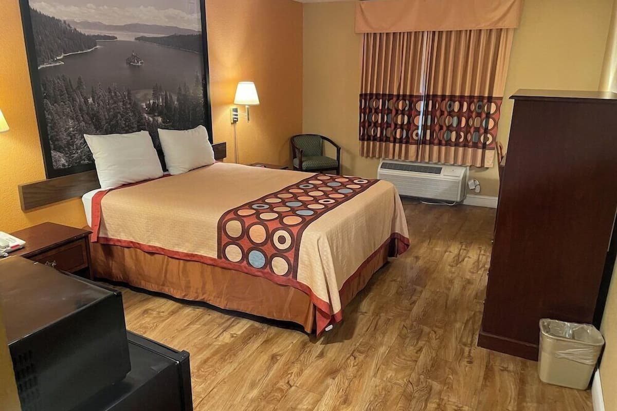A hotel room with a bed, a desk, a chair, a TV, a window with curtains, and a painting on the wall.
