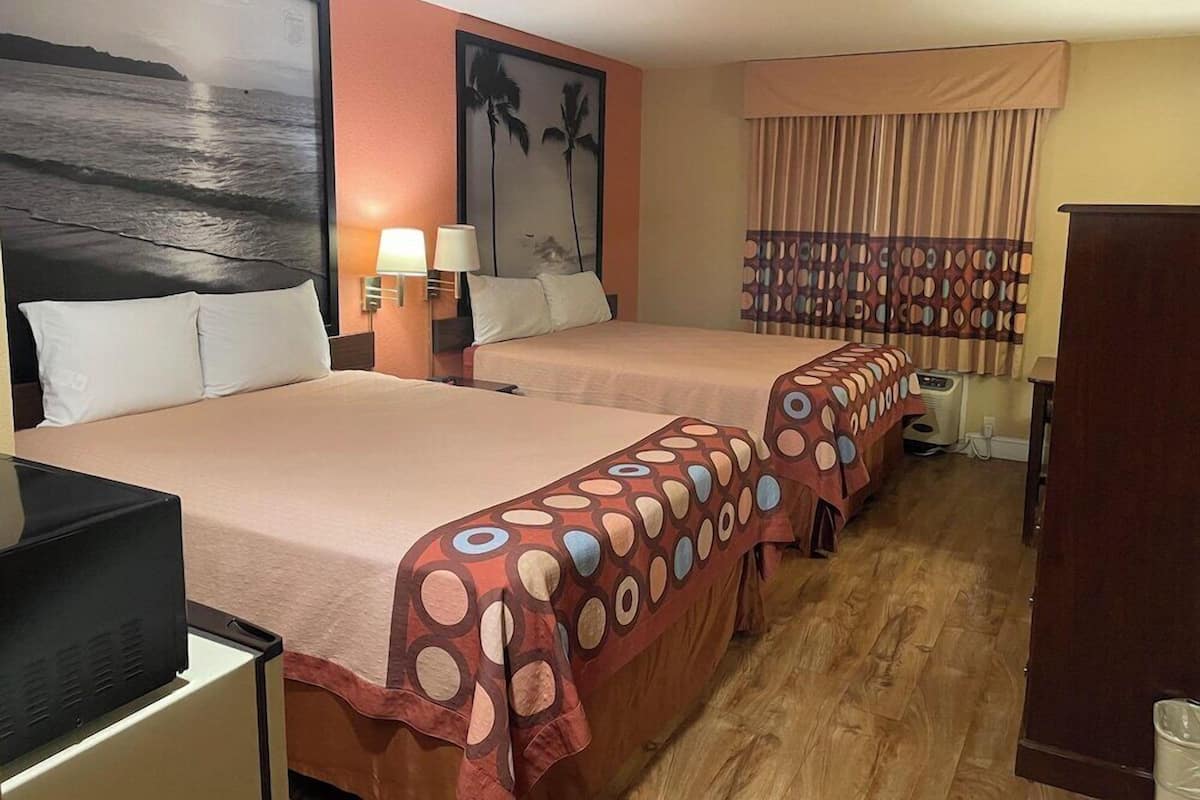 A hotel room with two beds, a desk with a computer, a wall-mounted TV, and a window with curtains.