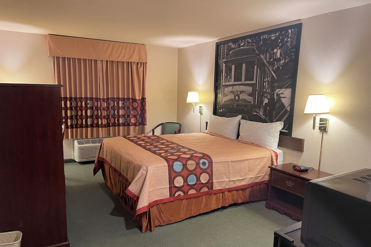 A hotel room with a bed, two bedside lamps, a desk with a computer, a TV, a chair, a window with curtains, and a wall-mounted picture.
