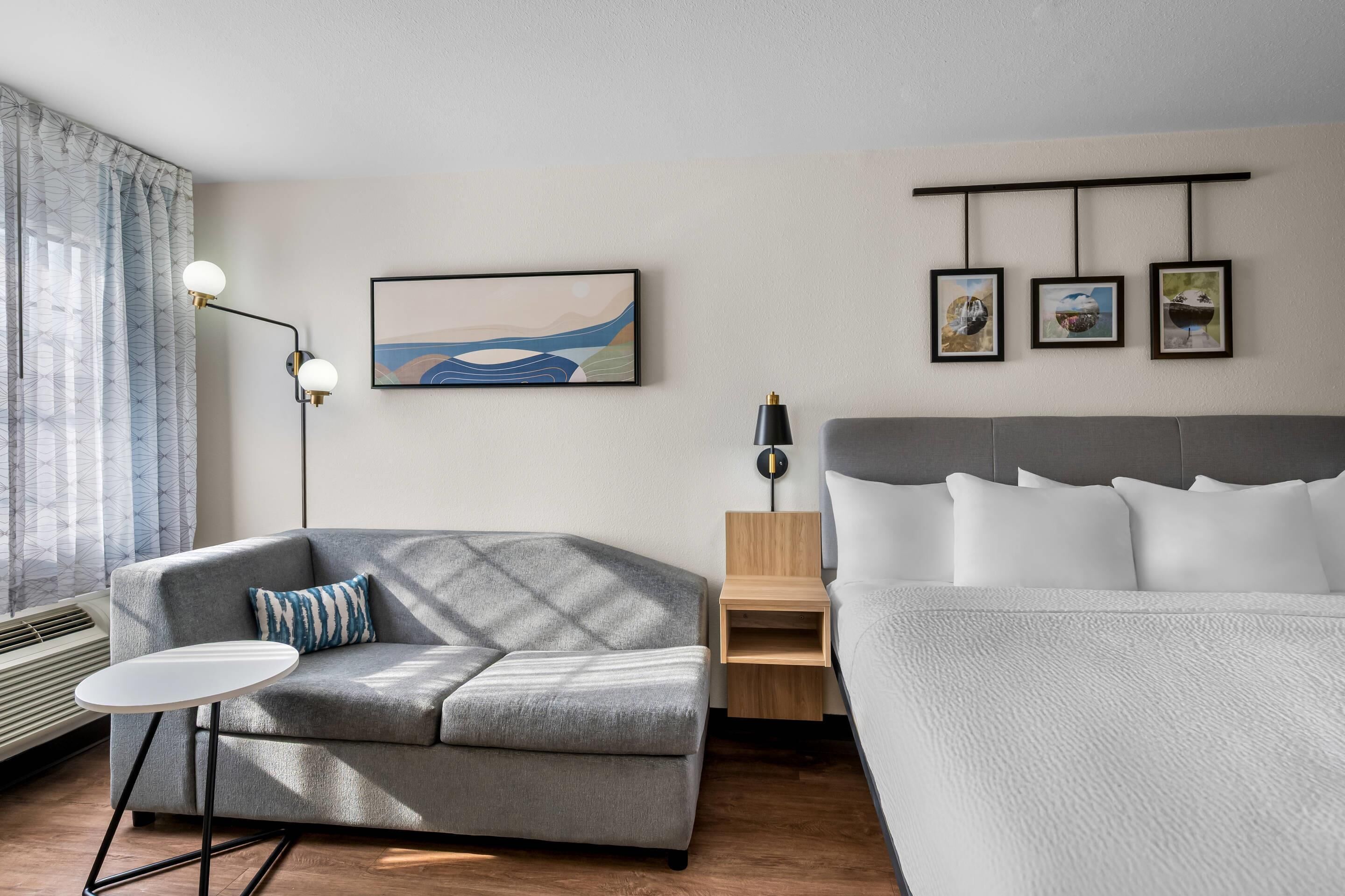 A modern hotel room with a grey sofa, a small round table, a bed with white bedding, a wall-mounted artwork, and a bedside table with a lamp.