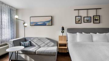 A modern hotel room with a grey sofa, a small round table, a bed with white bedding, a wall-mounted artwork, and a bedside table with a lamp.