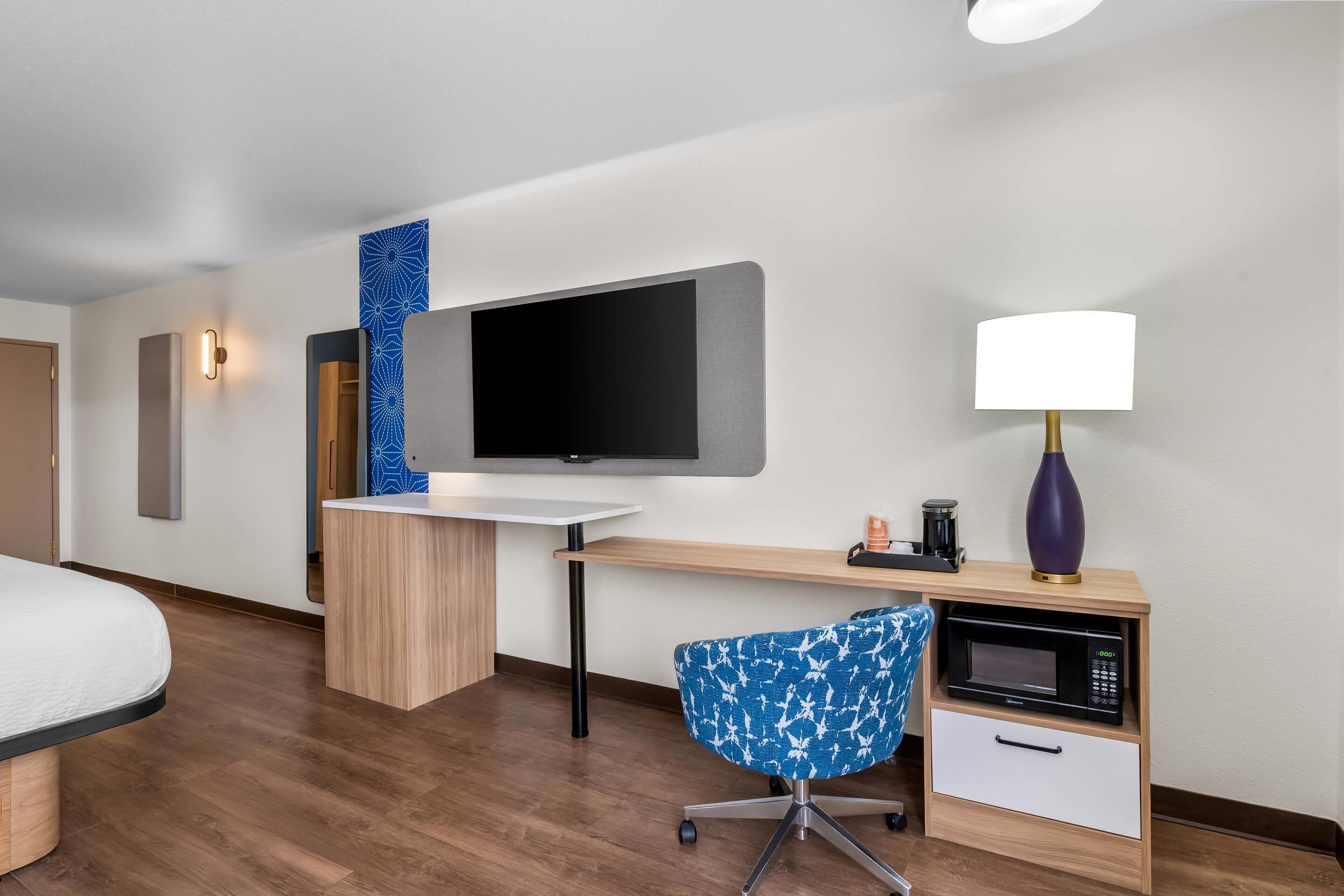 A hotel room with a bed, a desk with a flat-screen TV, a coffee maker, a lamp, a microwave, and a blue patterned chair.