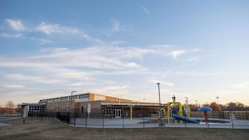 A playground with slides and swings, enclosed by a fence, and a building with a flat roof in the background.
