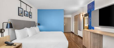 Una habitación de hotel moderna con una cama grande, una mesita de noche con lámpara, un escritorio con teléfono y un televisor montado en la pared.