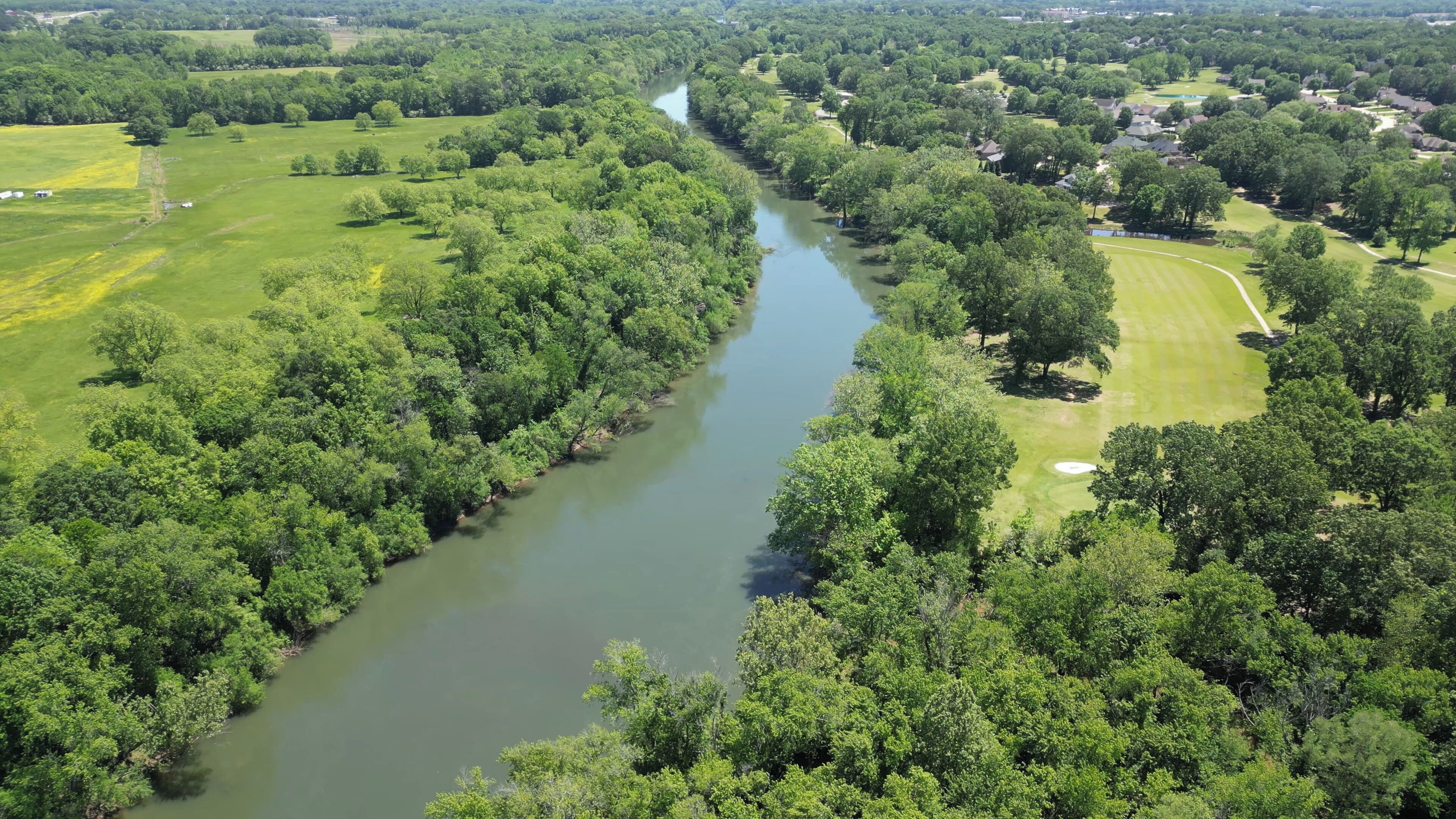 A river running through a densely wooded area with residential properties on one side and open fields on the other.