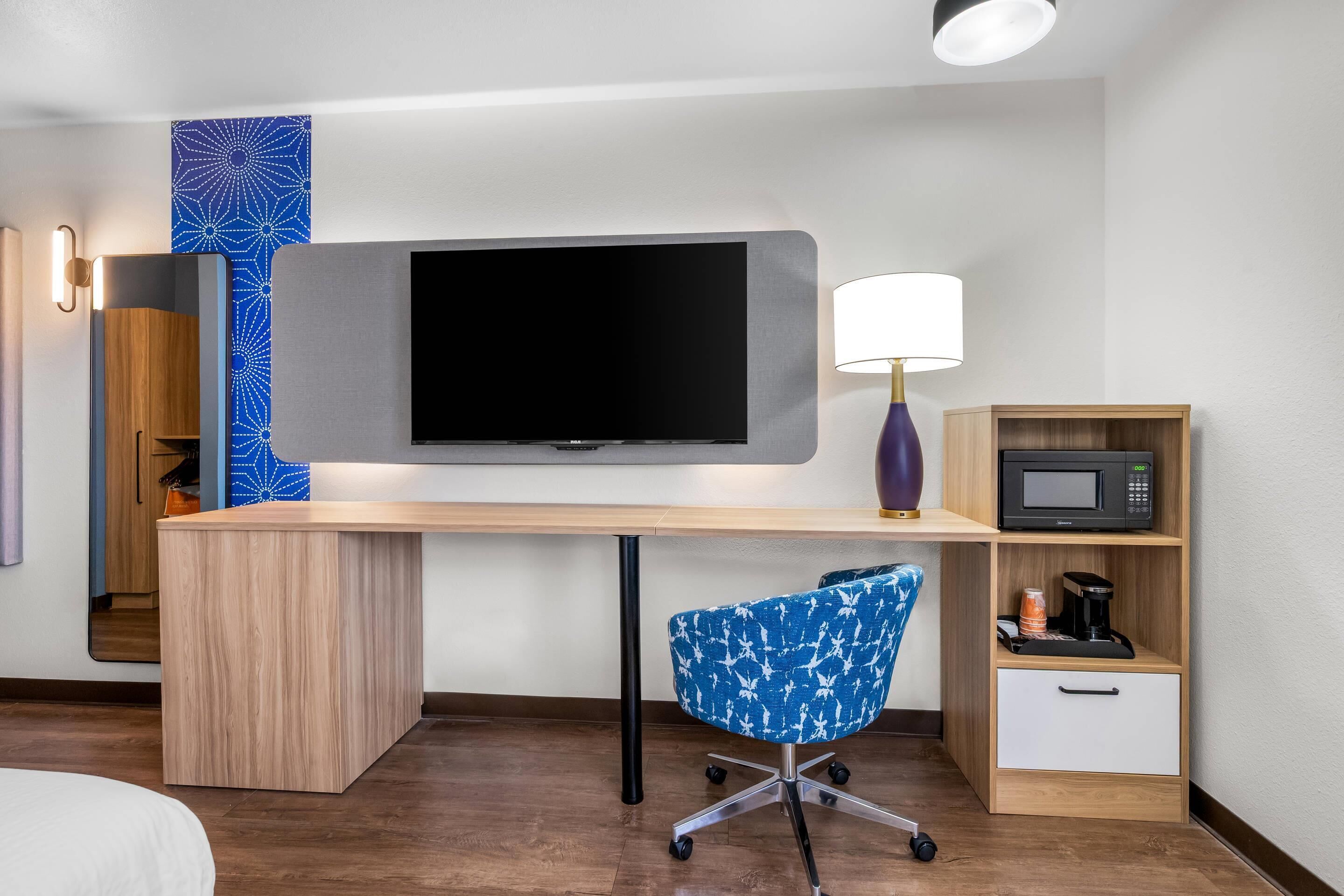 Quarto de hotel com escrivaninha de madeira, televisão de tela plana, cadeira de escritório azul, luminária, micro-ondas e máquina de café.