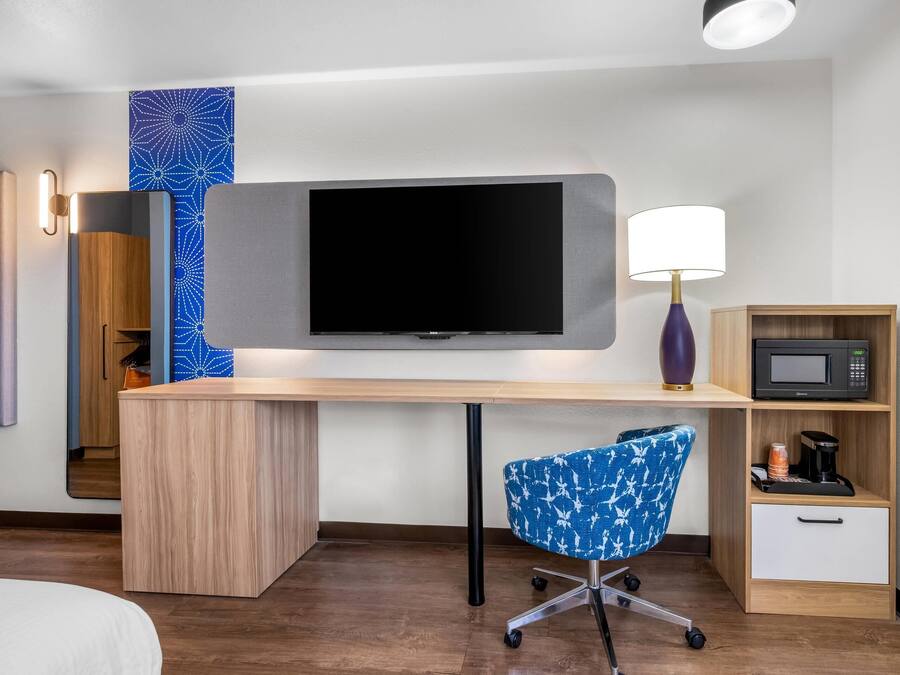 Habitación de hotel con un escritorio de madera, un televisor de pantalla plana, una silla de oficina azul, una lámpara, un microondas y una cafetera.