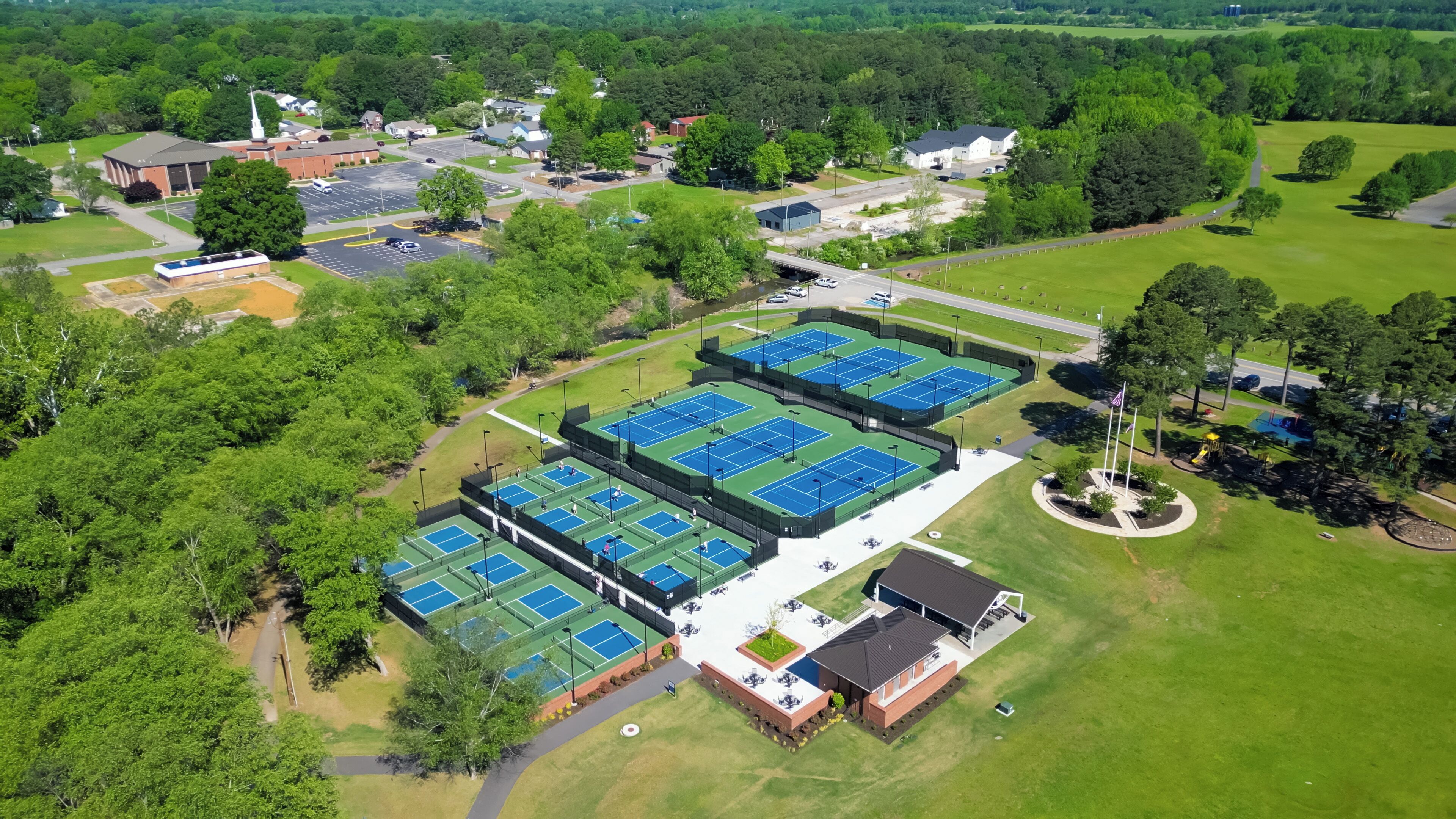 An aerial view of a sports complex with multiple tennis courts, a parking lot, and surrounding greenery.