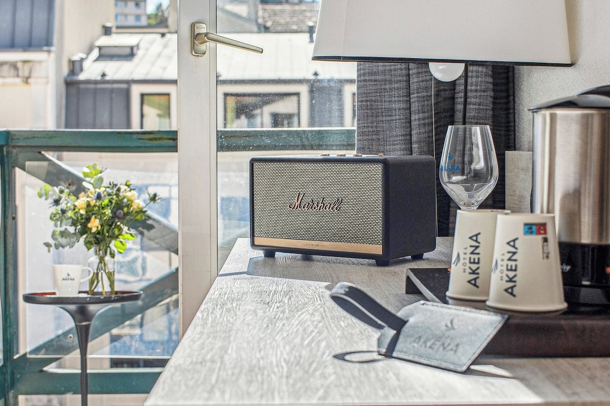 A hotel room with a view of buildings, a table with a lamp, a wine glass, and a minibar.