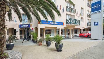 Ein Hotel namens 'AKENA' mit einer Schautafel, Pflanzgefäßen und einem roten Auto vor der Fassade.