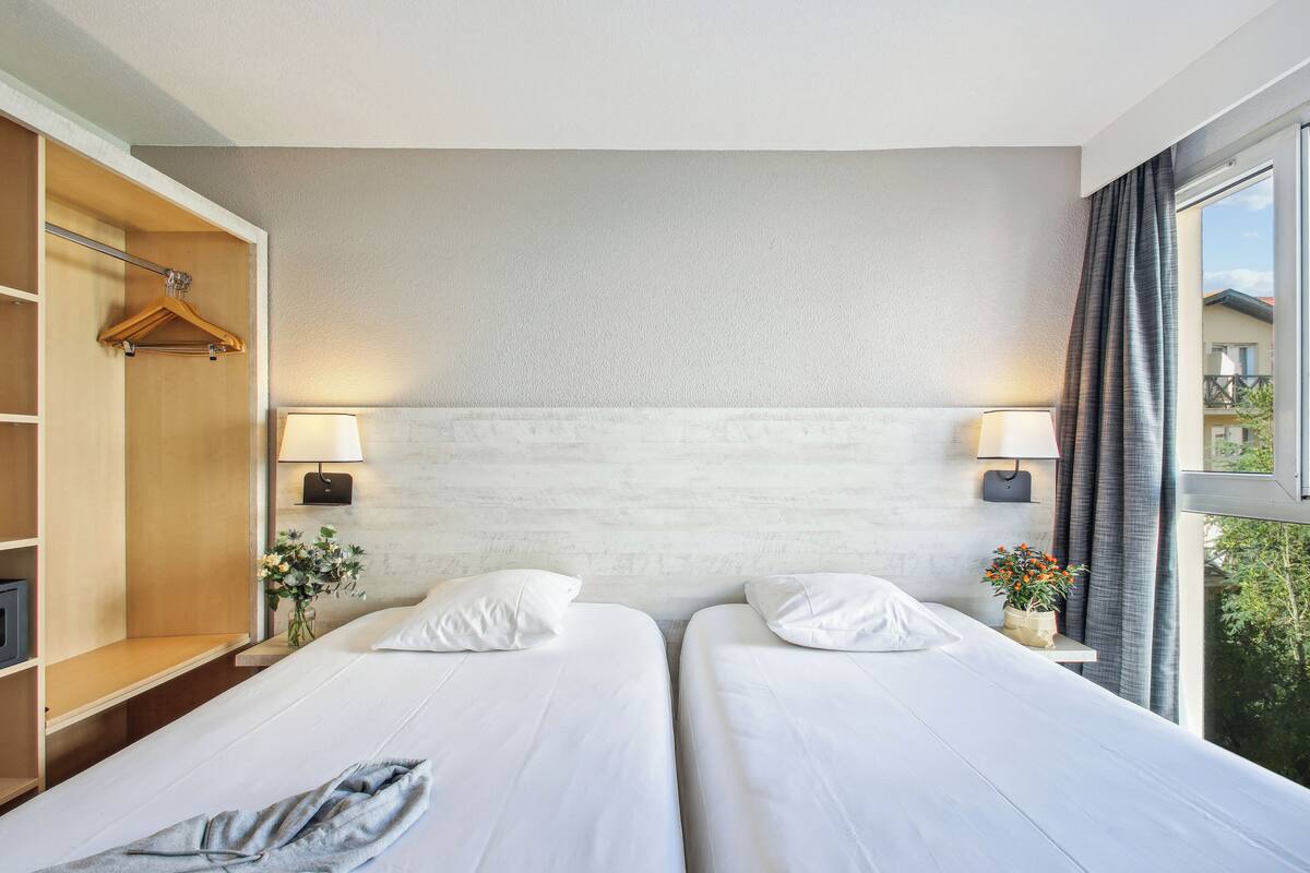 A hotel room with two beds, a wooden wardrobe, bedside lamps, and a window with curtains.