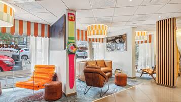 Un hall d’hôtel au design moderne, avec un canapé en cuir, un décor rayé orange et blanc, et une grande fenêtre donnant sur les voitures garées.