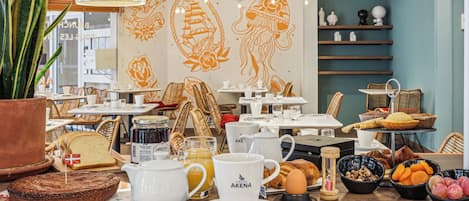 Mesa de café da manhã com diversas opções de alimentos, como pães, doces e frutas, dispostos sobre mesas com louças brancas.
