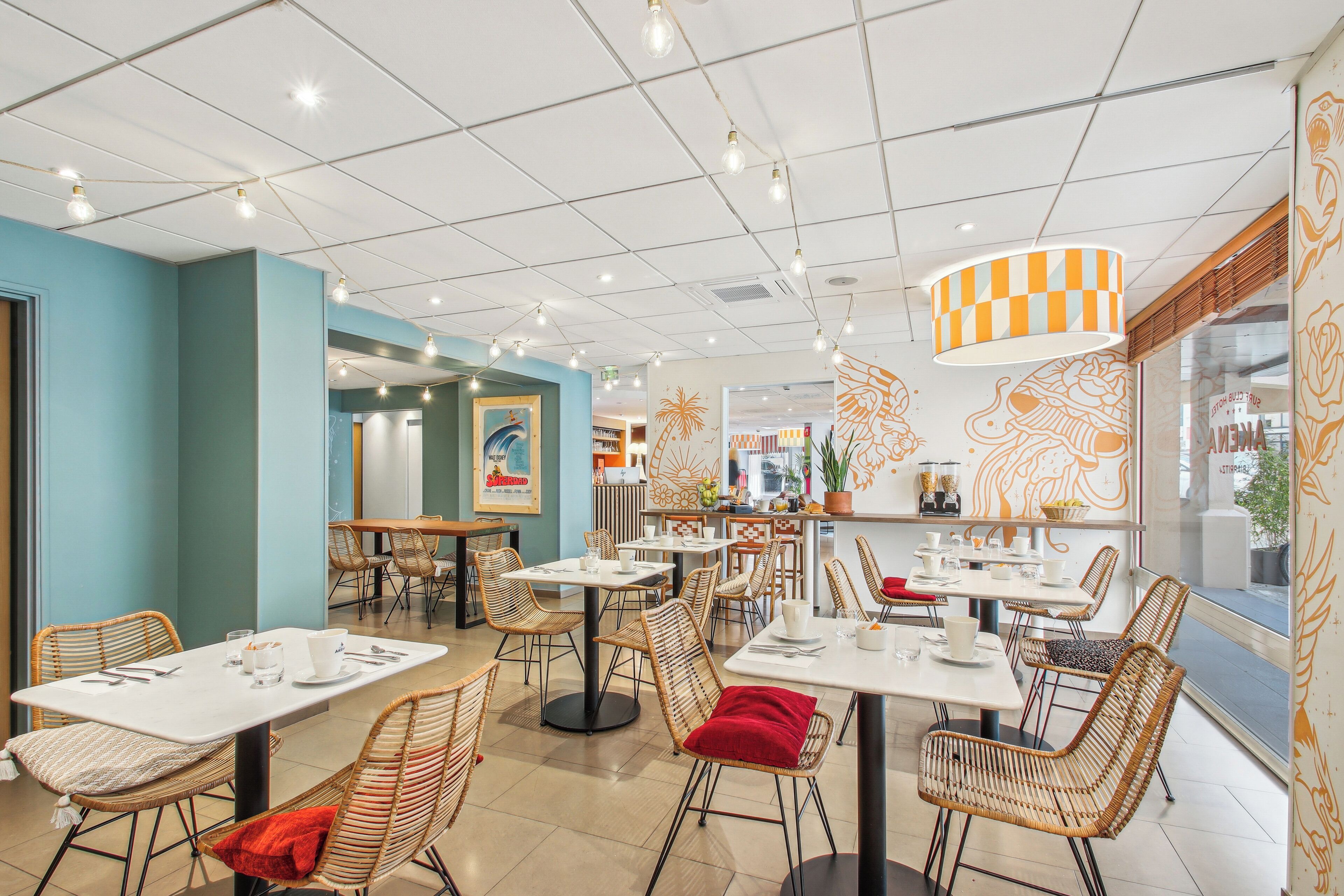 Un restaurant moderne doté de nombreuses tables et chaises, d’un luminaire au plafond à motifs à carreaux et d’œuvres d’art décoratives murales.