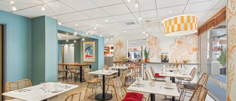 Un restaurant moderne doté de nombreuses tables et chaises, d’un luminaire au plafond à motifs à carreaux et d’œuvres d’art décoratives murales.