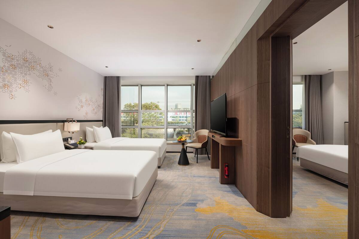 A hotel room with two beds, a TV, a desk, and a view of the city.