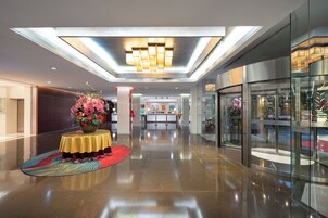 A modern hotel lobby with a polished floor, a central round table with a floral arrangement, and a glass elevator.
