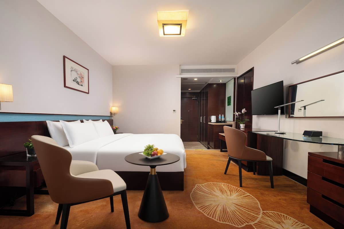 A hotel room with a bed, two chairs, a desk, a television, and a small table with a fruit bowl.