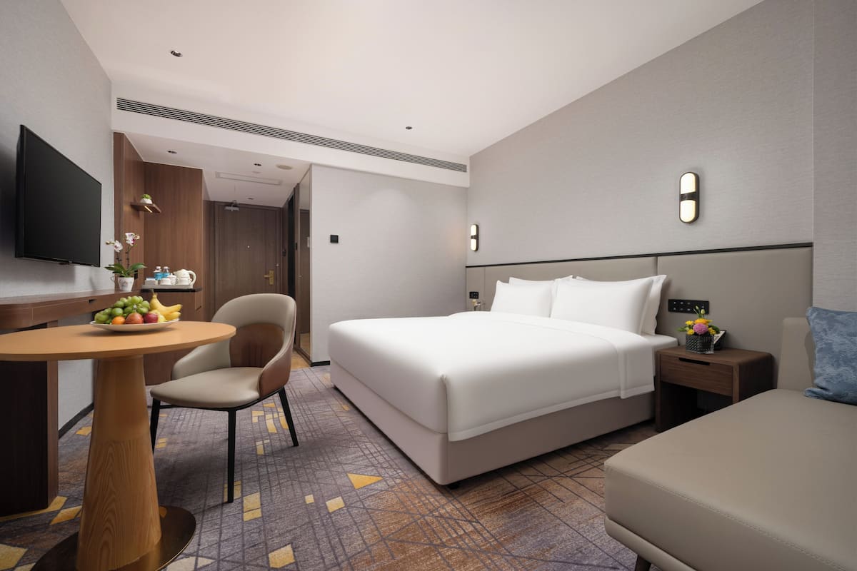 A hotel room with a bed, a desk with a chair, a TV, and a small table with fruit.