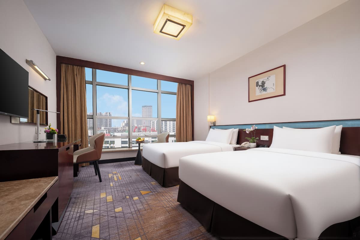 A hotel room with two beds, a desk, a chair, a TV, and a large window with a city view.