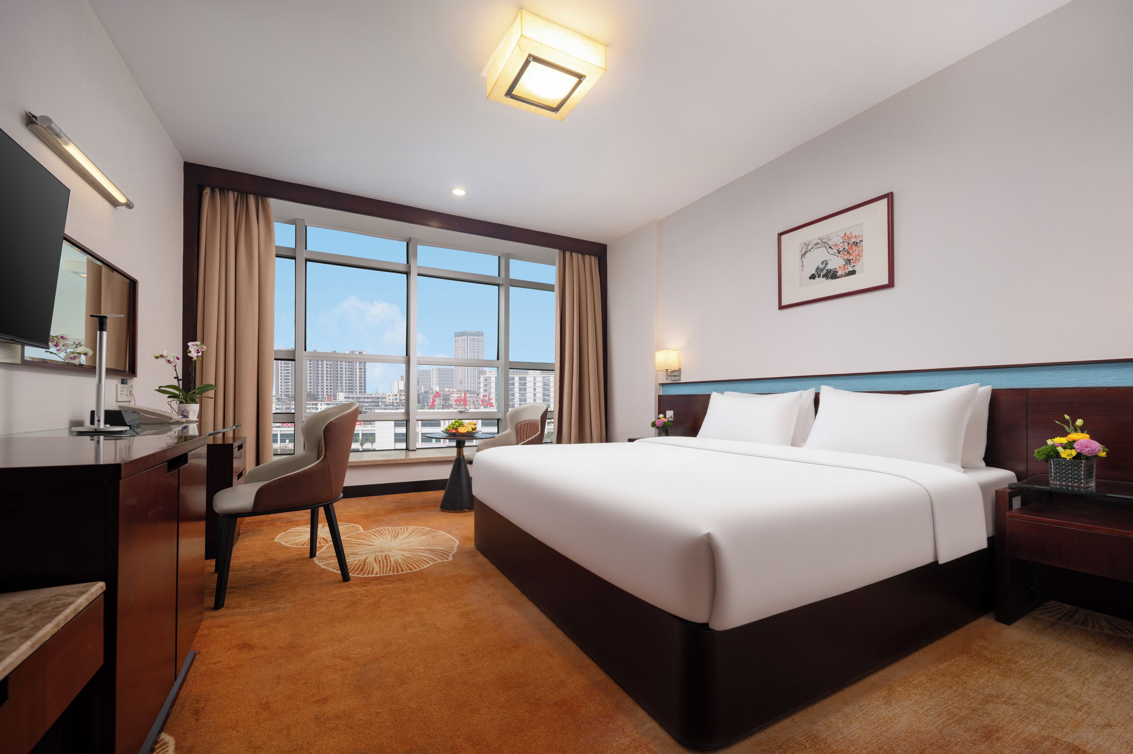 Quarto de hotel com uma cama grande, uma escrivaninha, uma cadeira, uma mesa pequena, vista da cidade e um quadro na parede.