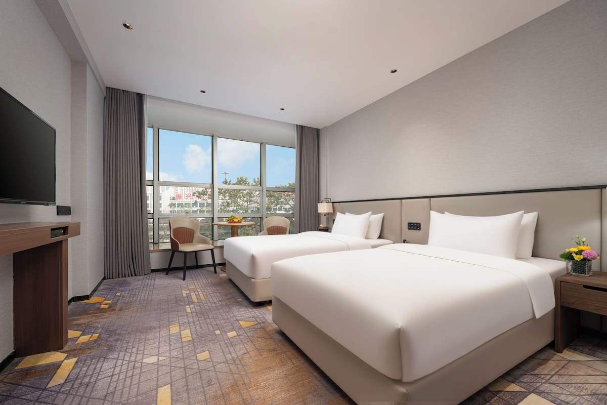 A hotel room with two beds, a desk, a TV, and a large window with a city view.