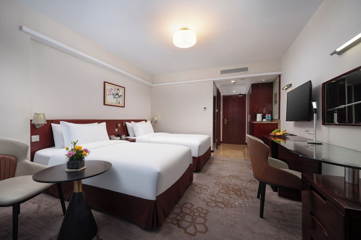 A hotel room with two beds, a desk, a chair, a TV, and a small table with flowers.
