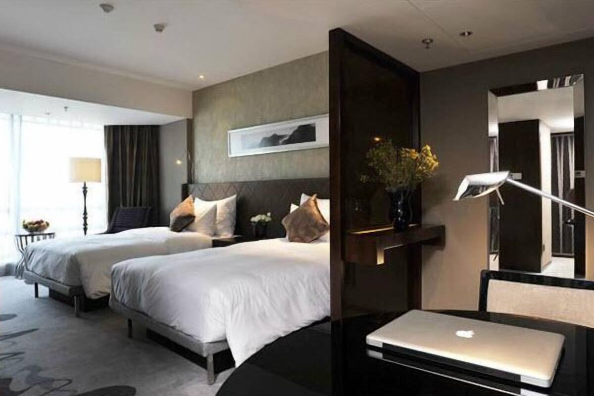 A hotel room with two beds, a desk with a laptop, a lamp, and a vase with flowers.
